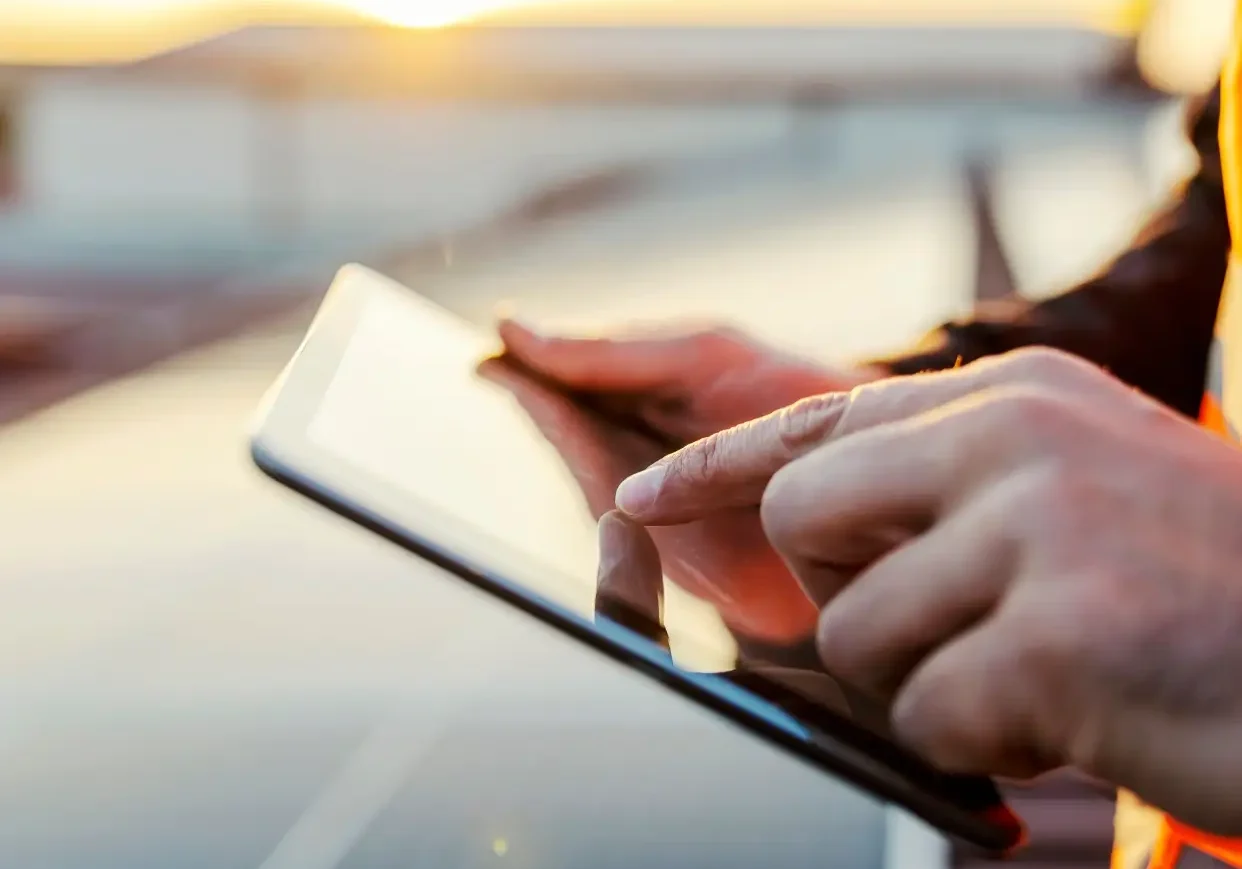 Close-up of a person using a digital tablet outdoors, with solar panels and a setting sun in the background.